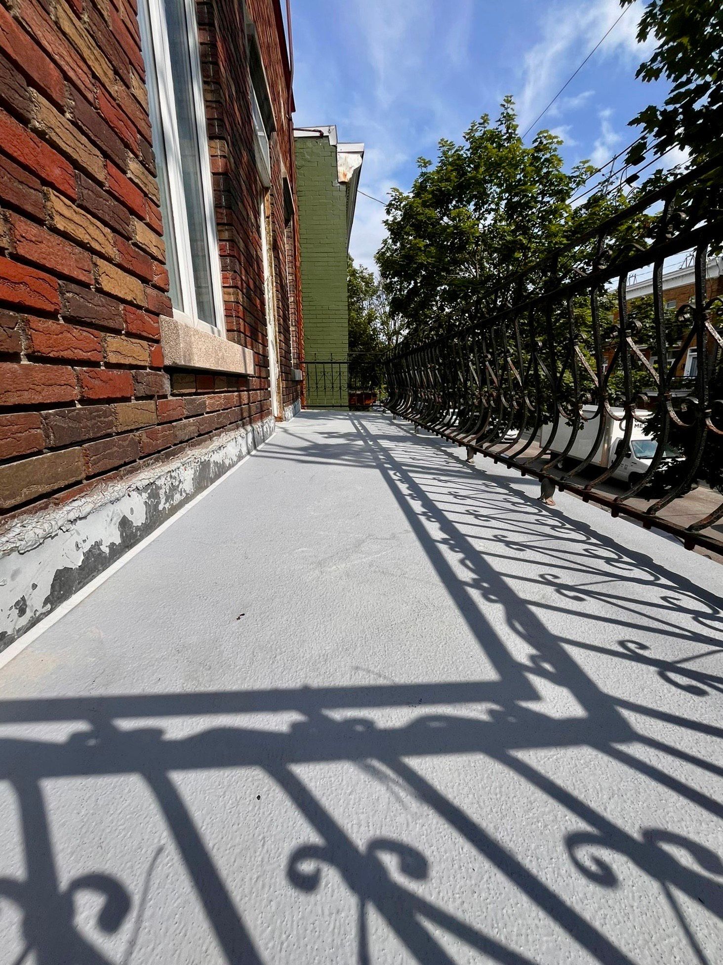 Vue en contre-plongée d'un sol de balcon gris fraîchement recouvert, adossé à un mur de briques, avec l'ombre de la rambarde projetée sur la terrasse.