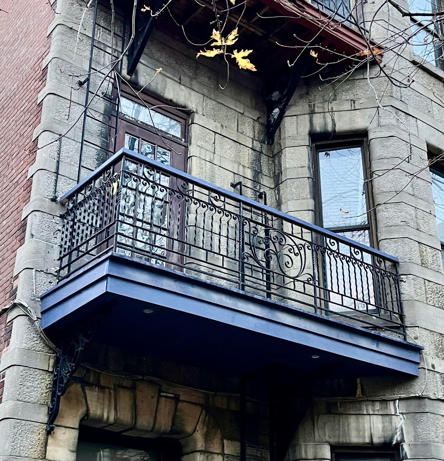 Façade en pierre avec balcon et porte en métal foncé. Balcon peint en bleu avec balustrade décorative.