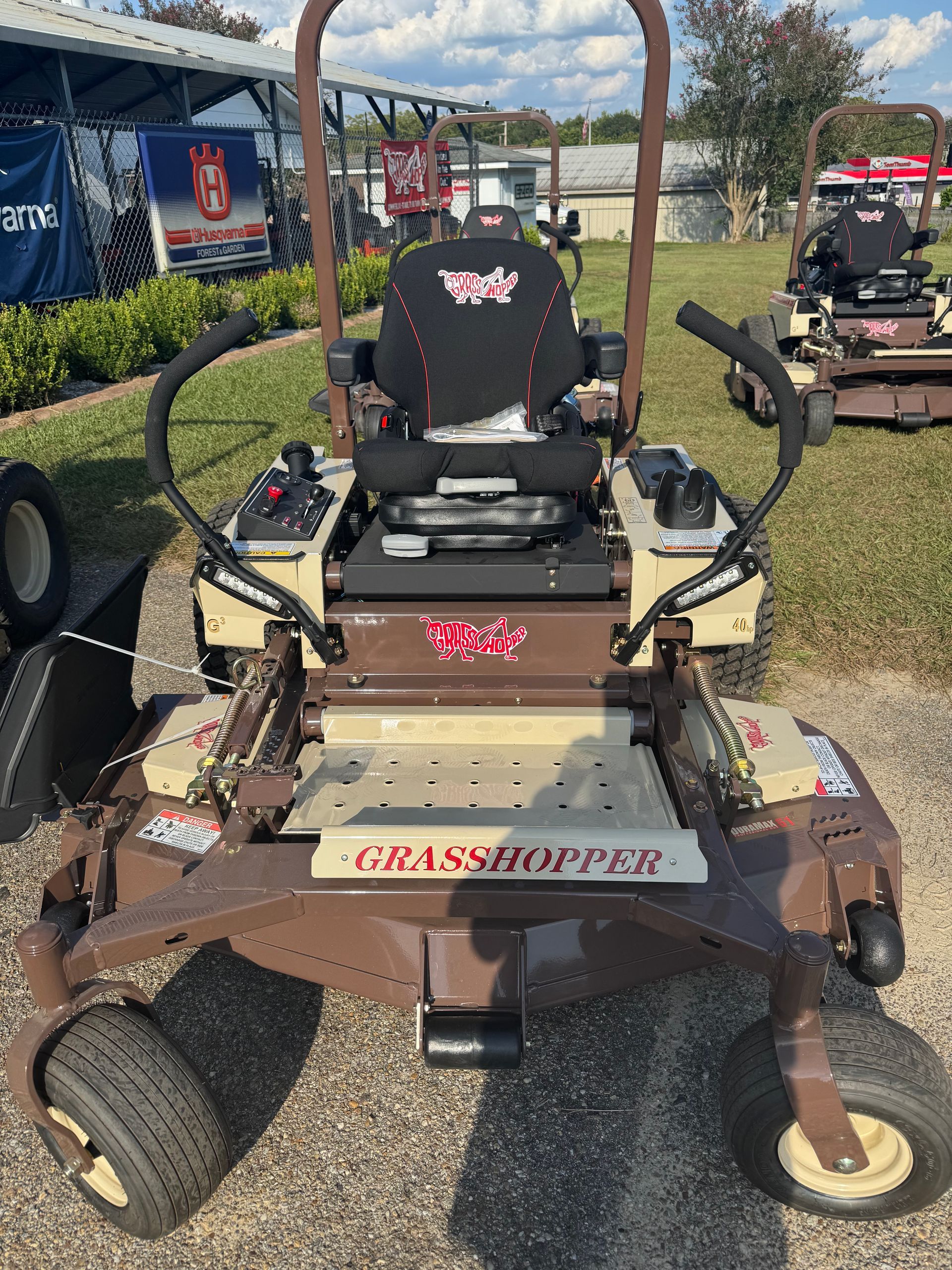 Brown Grasshopper zero-turn lawnmower on pavement, outdoors. Features seat, controls, and mower deck.