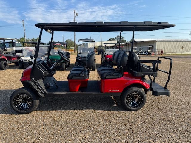 Red six-seater golf cart on gravel, with black roof, seats, and wheels.  Other golf carts in the background.