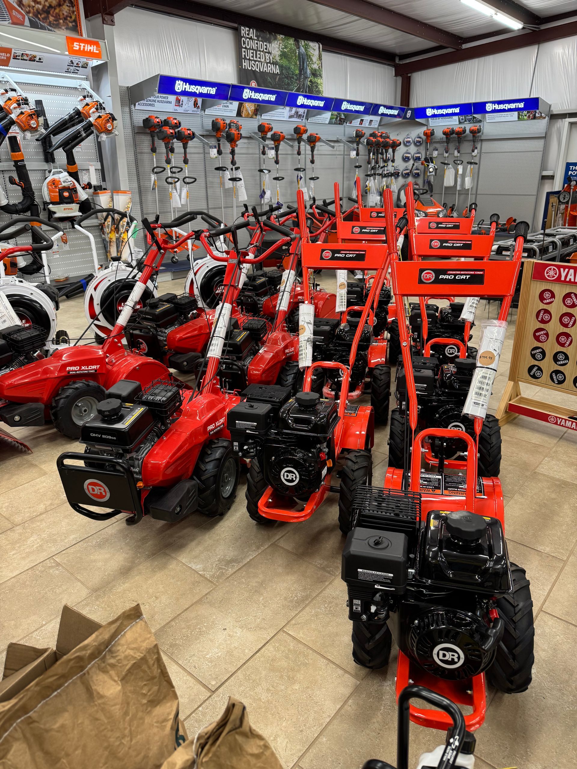 A store aisle featuring rows of red and black tillers, with other landscaping equipment in the background.