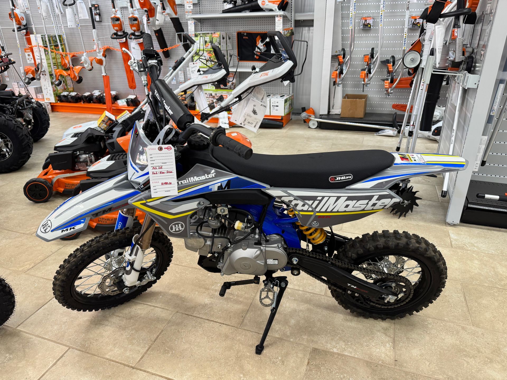 Dirt bike, blue and white TrailMaster brand, on display in a store, with other equipment in the background.
