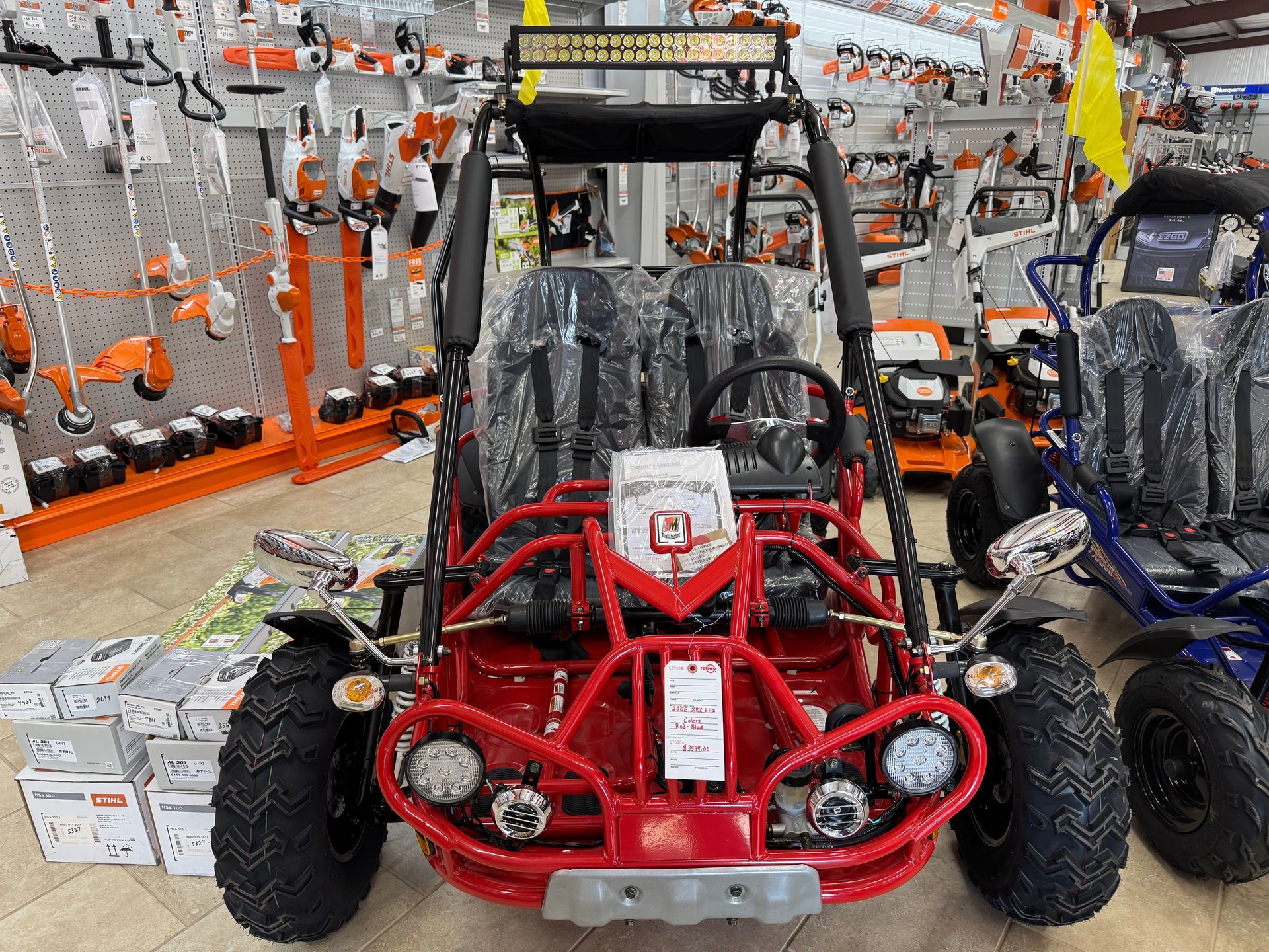 Red go-kart with black tires and roll cage on display in a store, surrounded by yard equipment.