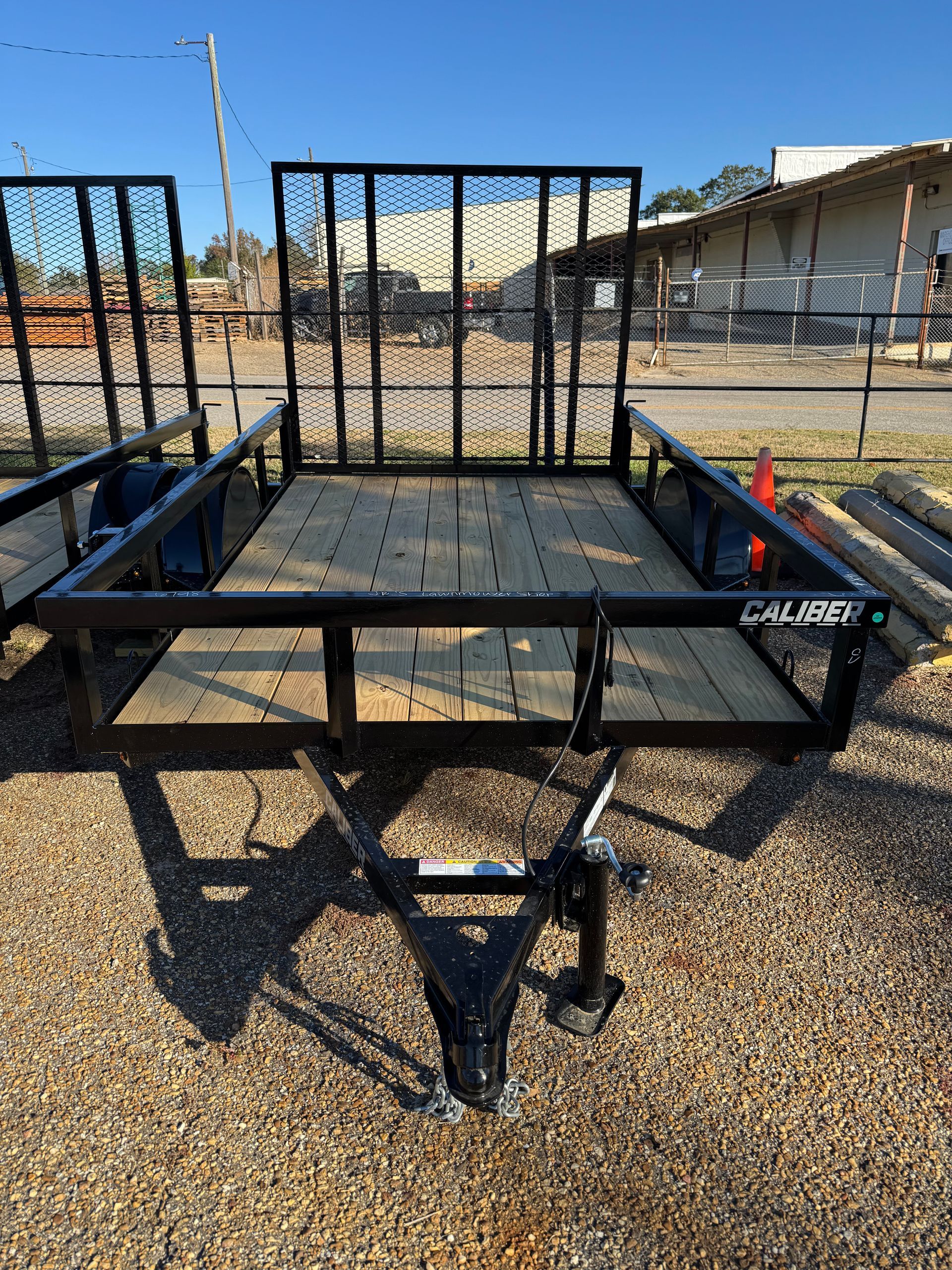 Black Caliber utility trailer with wood bed and wire mesh sides, parked outside on gravel.