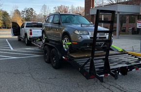 Gray SUV loaded on a black car hauler trailer in a parking lot, with a white pickup behind it. | Retana's Auto Repair LLC