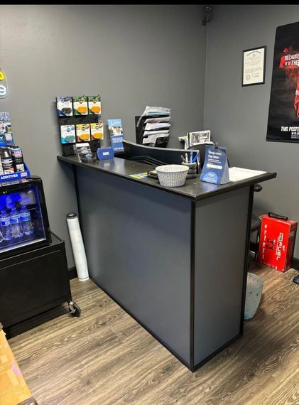 Small office reception desk with brochures, countertop items, and a mini fridge against gray walls | Retana's Auto Repair LLC