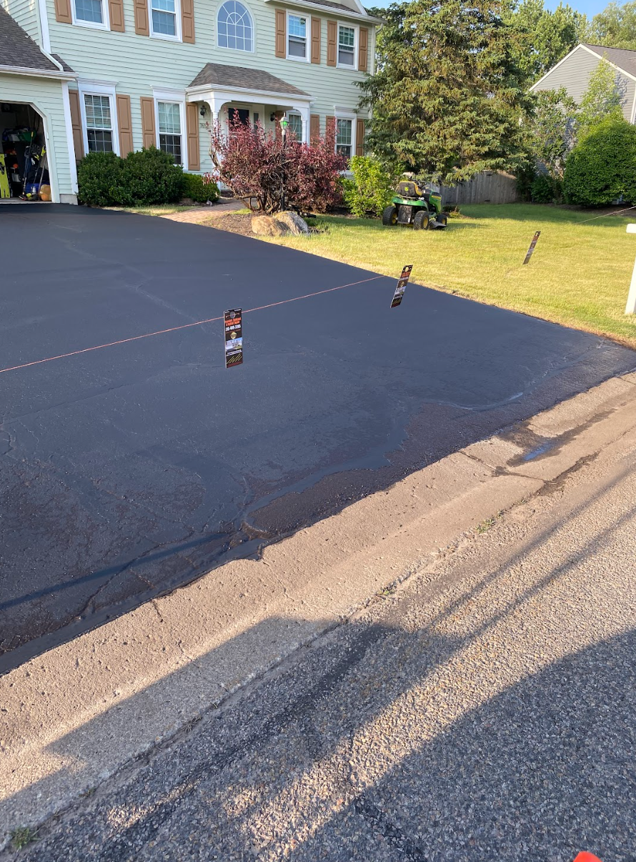 A skateboard is sitting on the side of a asphalt driveway in front of a house.