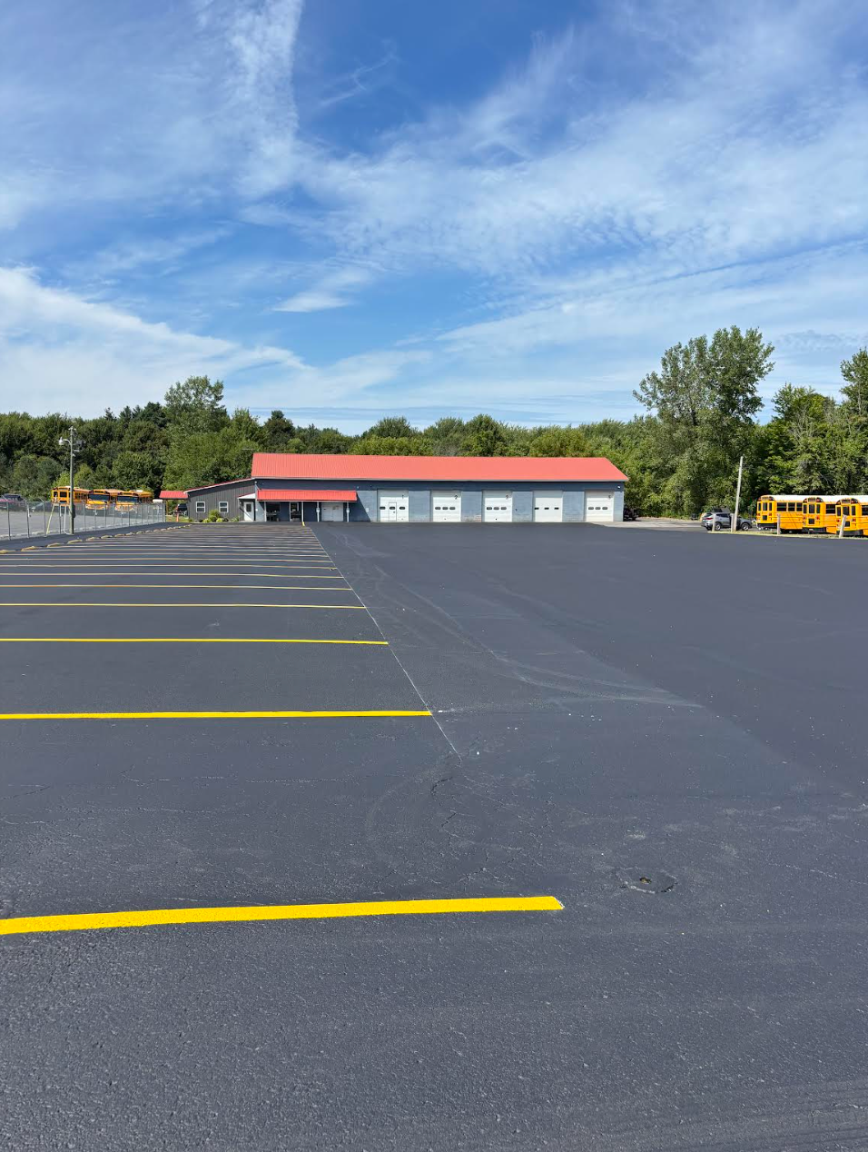 Empty parking lot with yellow lines leading to a building with a red roof and three garage doors. Blue sky.