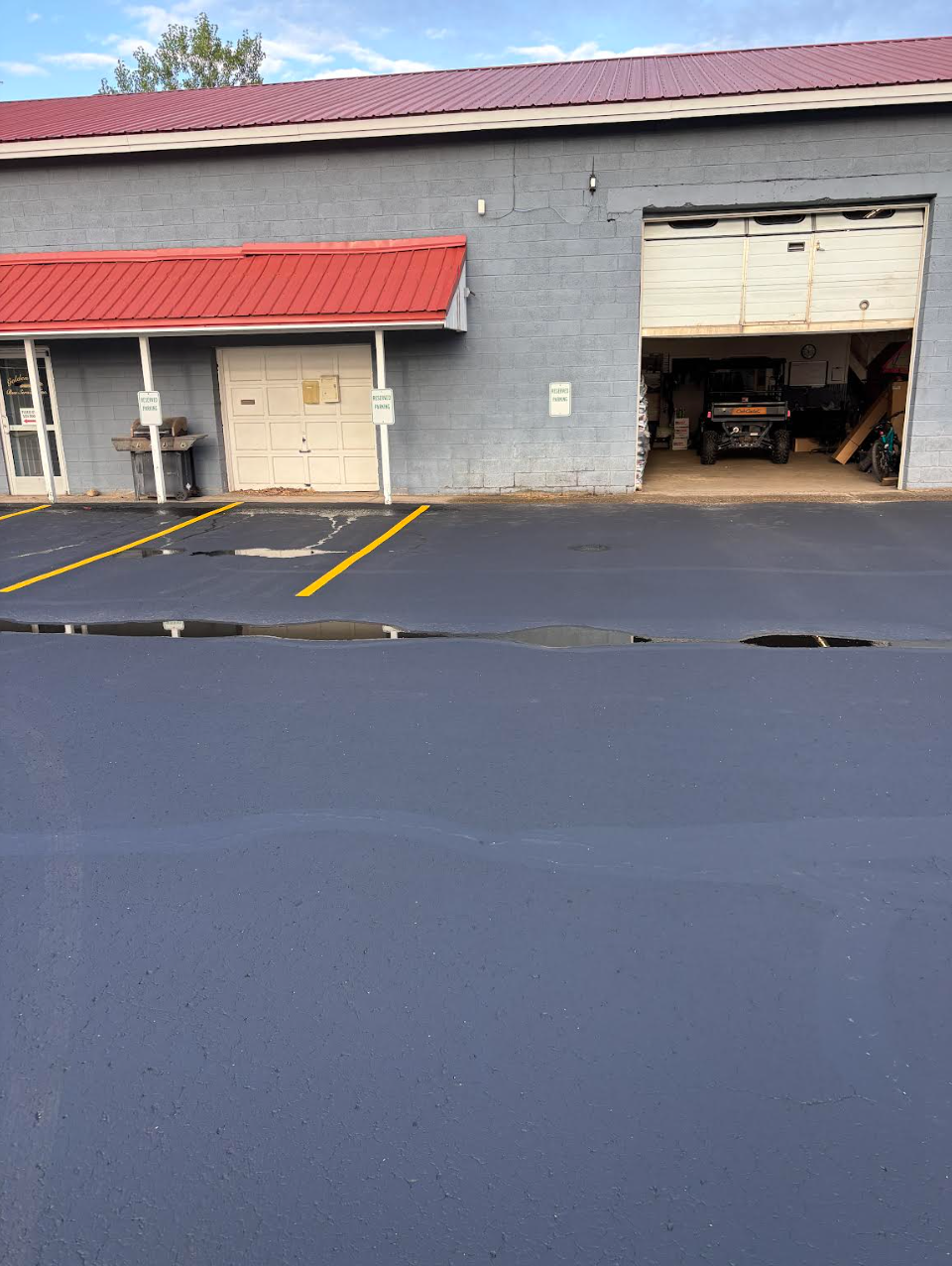 Gray commercial building with red roof and open garage door. Paved parking lot with yellow lines.