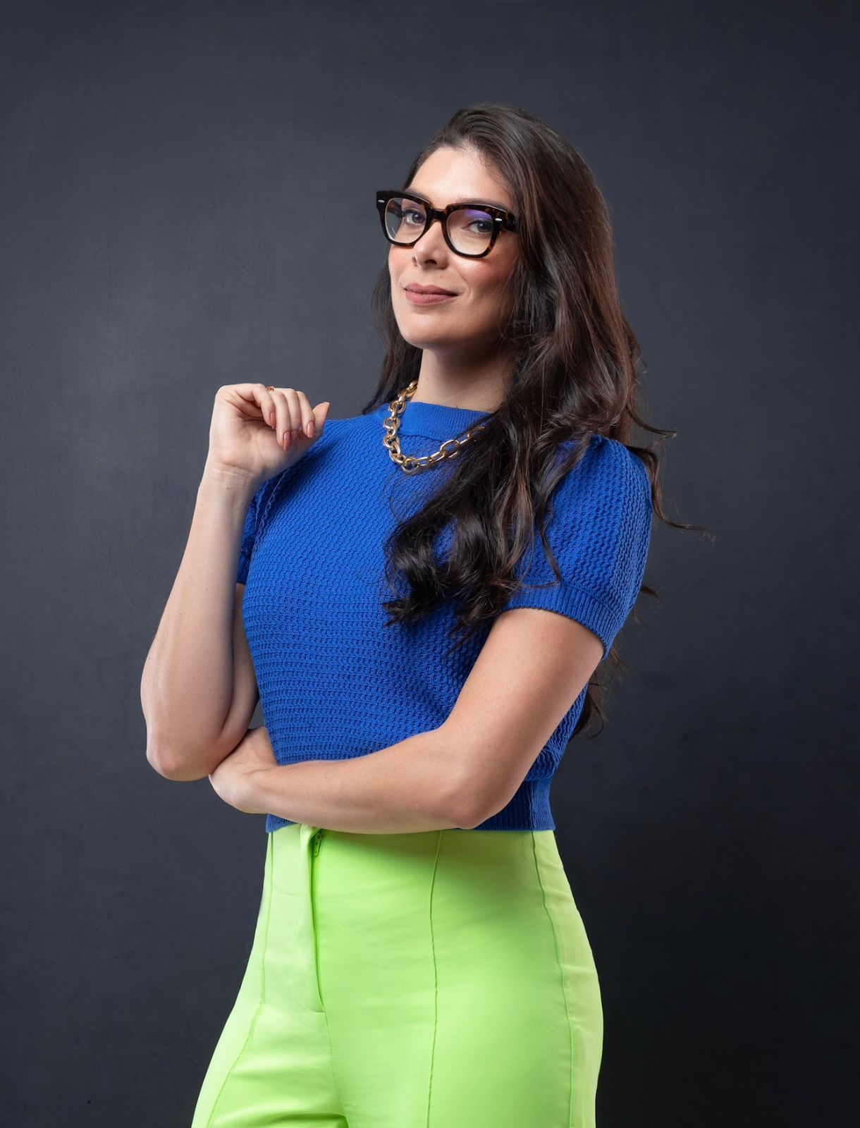 Uma mulher de óculos e camisa azul está de pé com os braços cruzados.