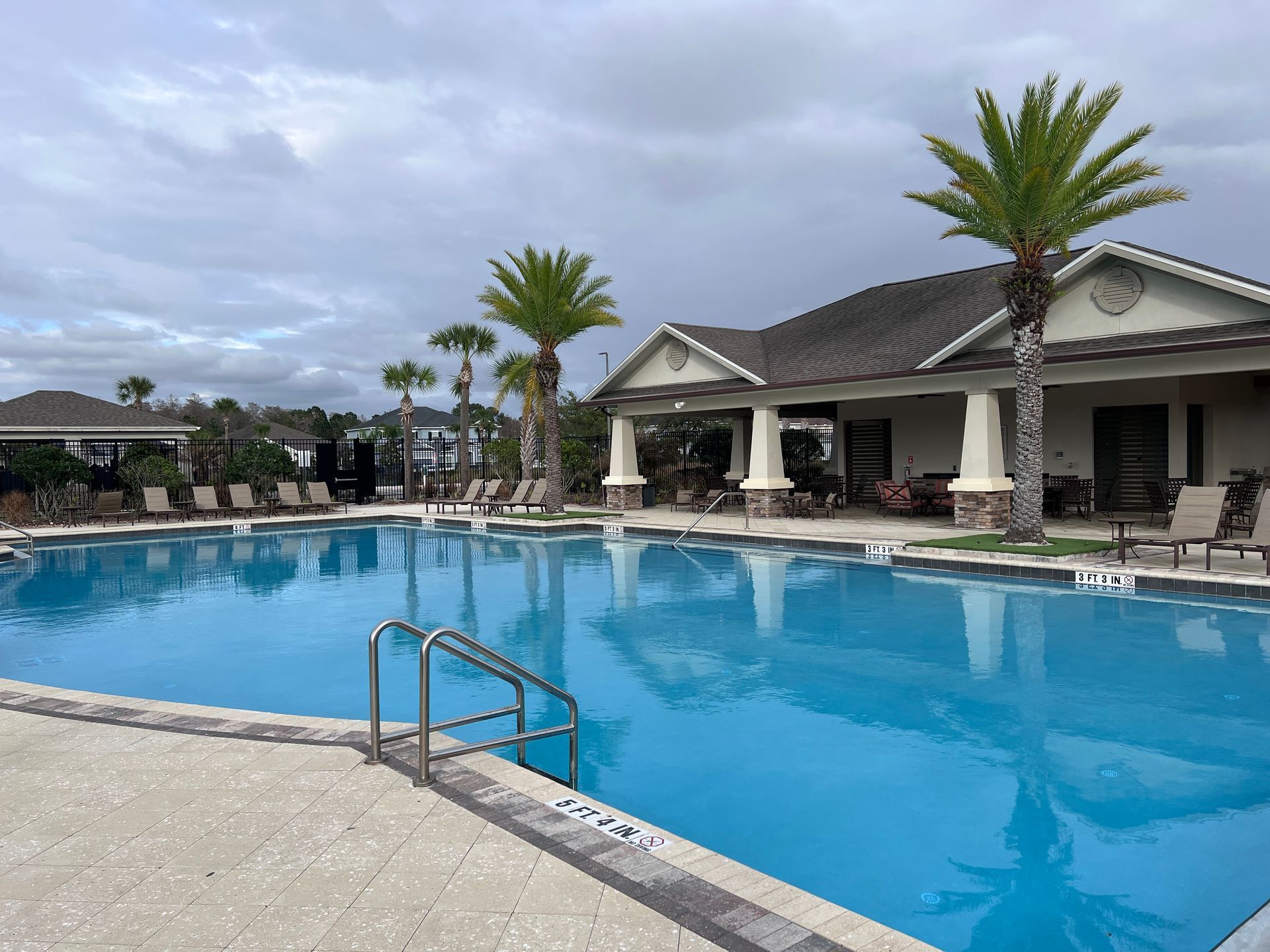 Outdoor resort pool with clear blue water, palm trees, and a covered patio under cloudy skies