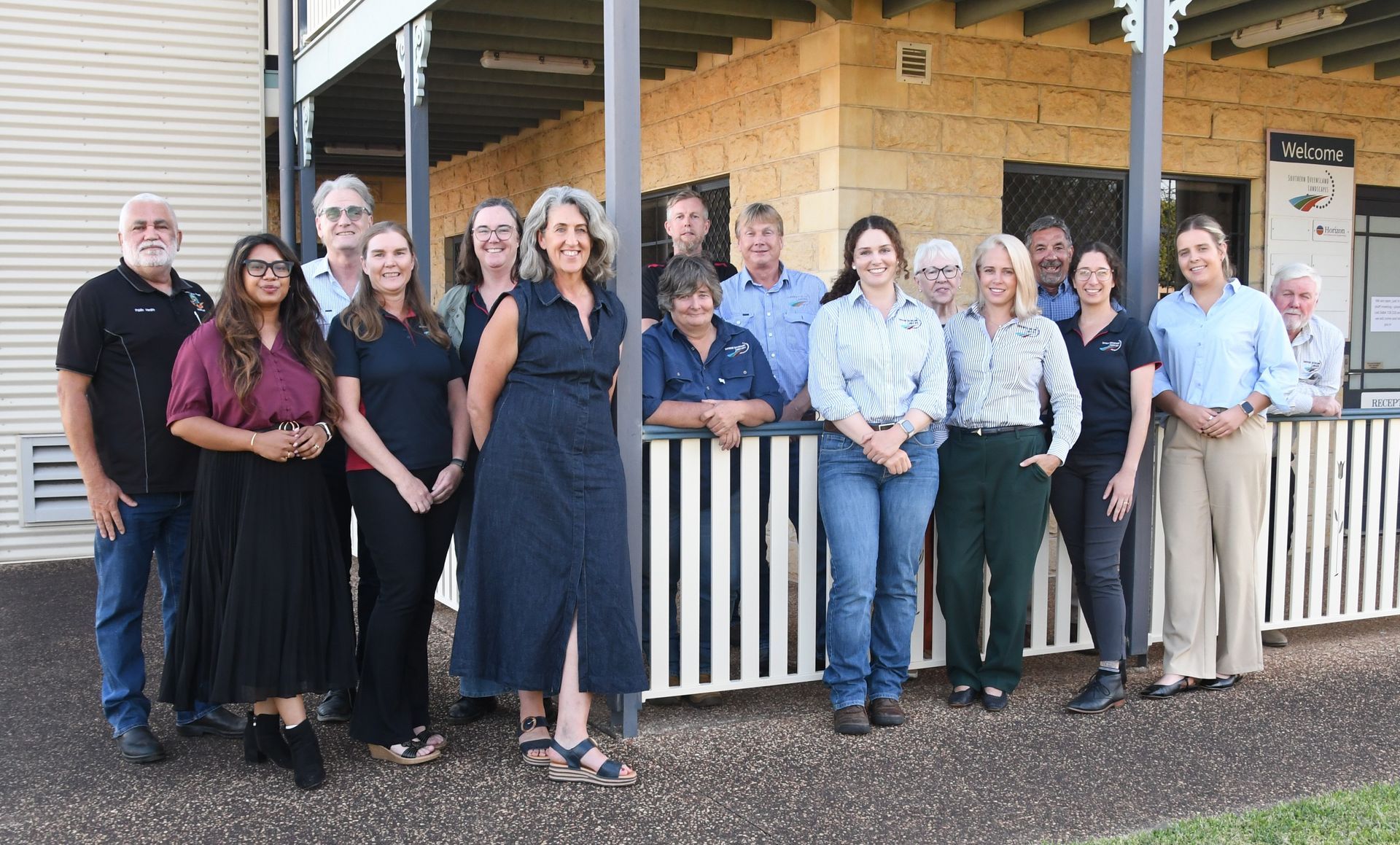 Group of people standing outside a building on a porch, posing together and smiling.