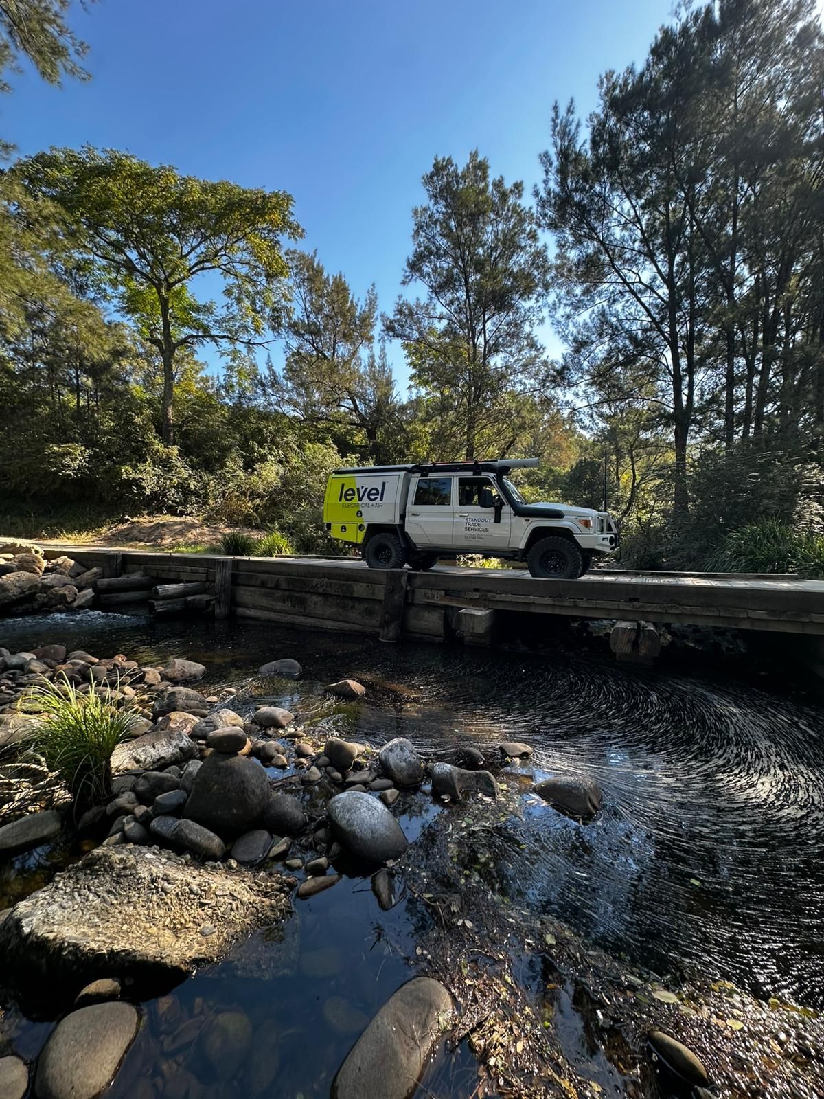 White off-road SUV crossing a shallow creek on a wooden bridge in a wooded area  — Level Electrical & Air In Taminda, NSW