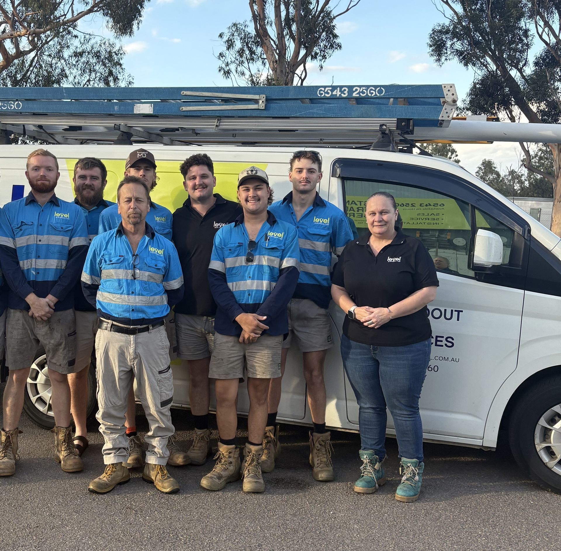 Seven utility workers in blue shirts standing beside a service van and ladder outdoors — Level Electrical & Air In Muswellbrook, NSW