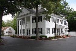 Large white colonial-style house with black shutters and columns, surrounded by trees and a paved driveway