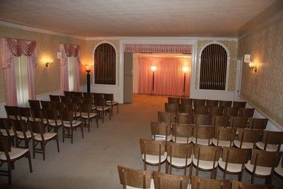 Empty banquet hall with rows of chairs facing a small lit stage and curtained windows.