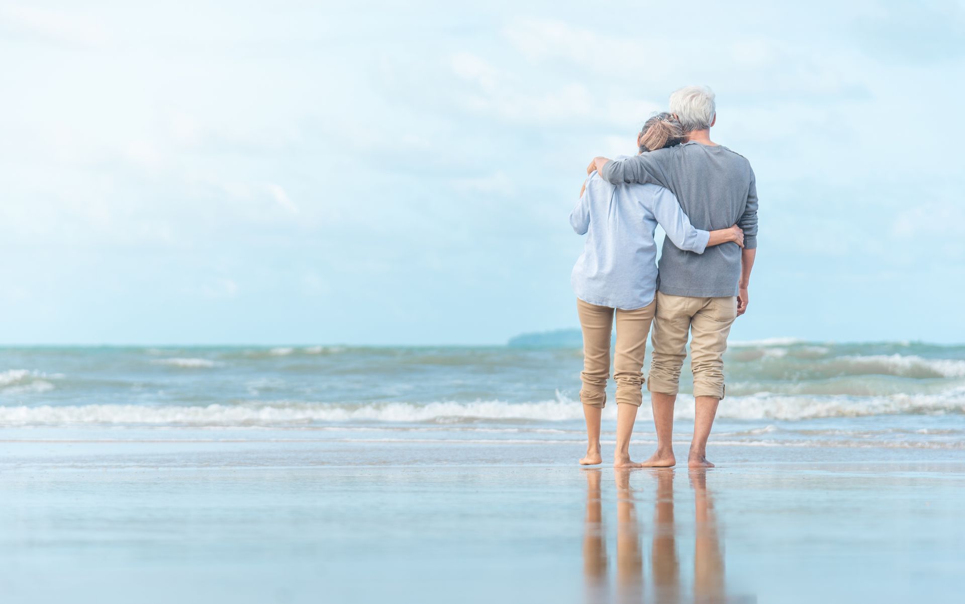 Two people standing arm-in-arm on a beach, facing the calm ocean under a bright sky.