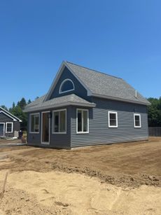 Small gray house under construction with white trim and unfinished dirt yard under a blue sky