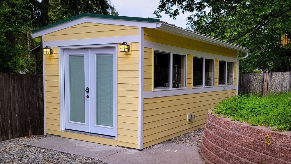 Yellow garden shed with white trim, double doors, and side windows beside a paved path and brick planter