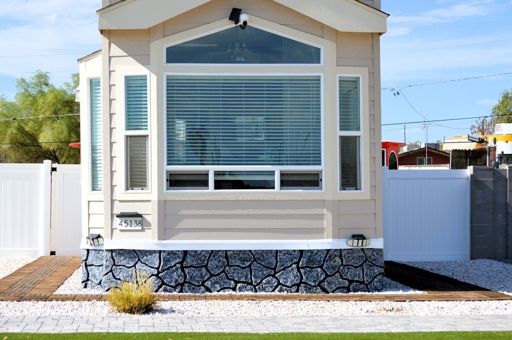 Small beige house with white trim, large front window, stone foundation, and white fence on a sunny day