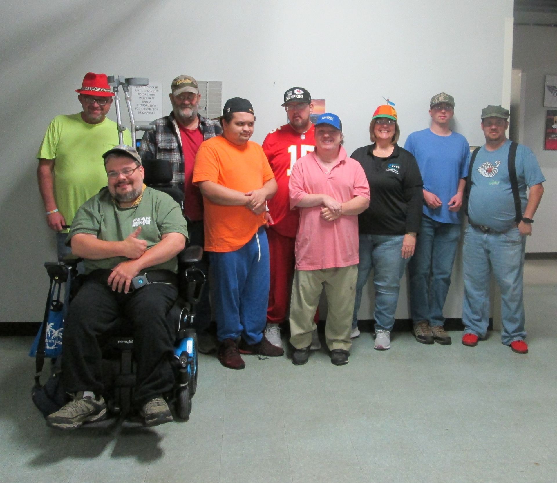 Group of people indoors, some wearing hats, smiling. One person in a wheelchair. Group of people indoors, some wearing hats, smiling. One person in a wheelchair.