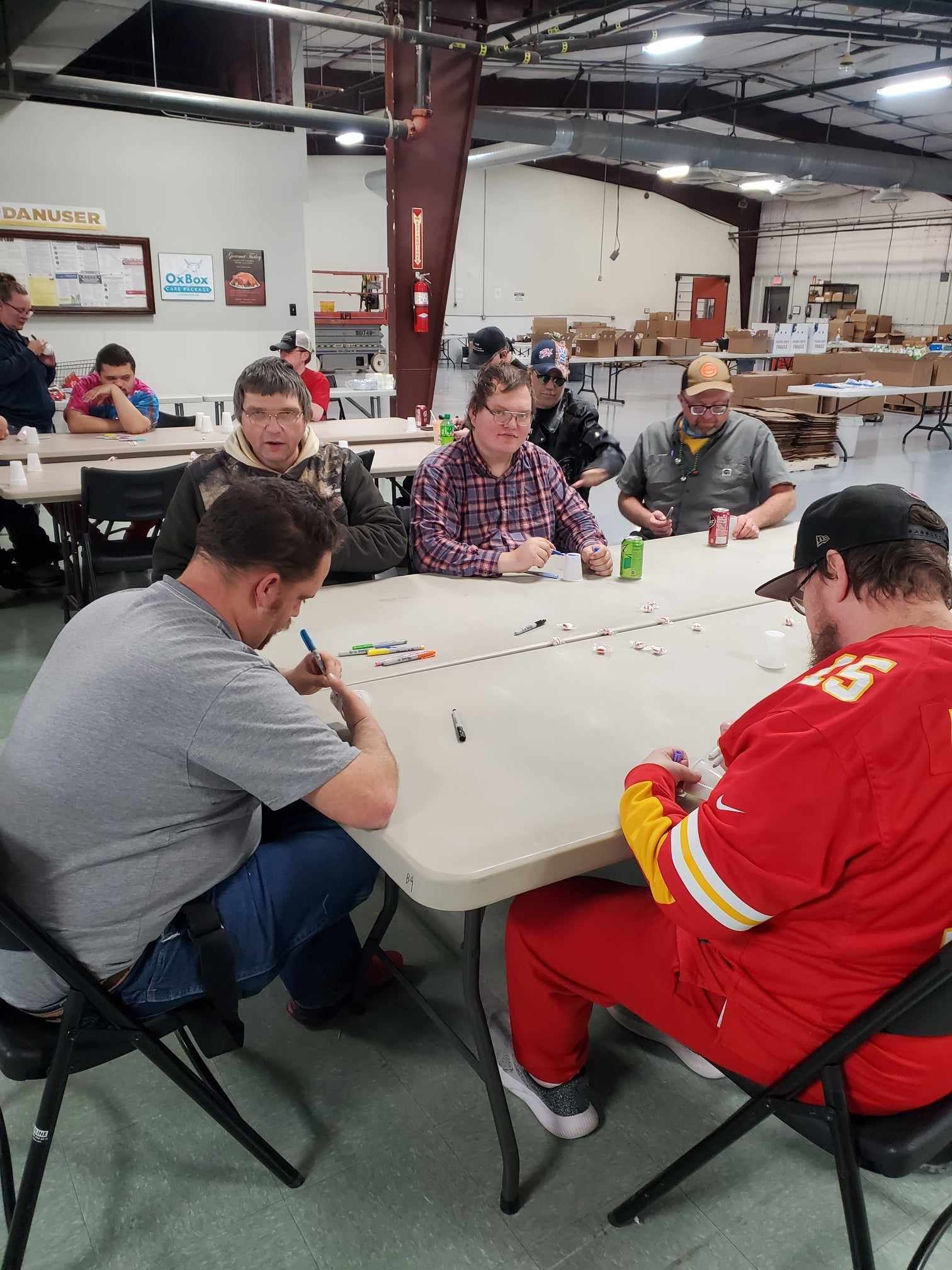 People sitting around a table, playing a game in an indoor setting. Some are writing, with drinks nearby. People sitting around a table, playing a game in an indoor setting. Some are writing, with drinks nearby.