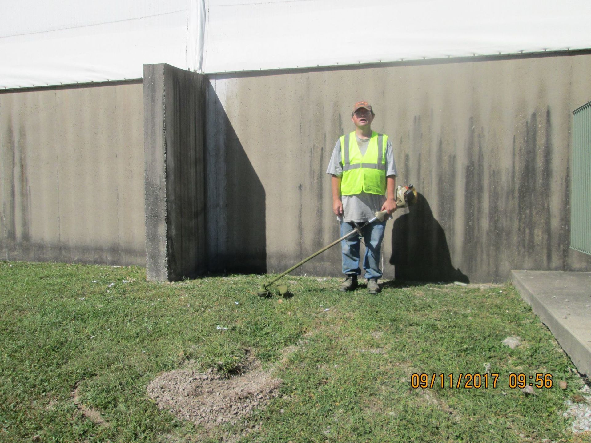 Person in safety vest trimming grass near a building with a weed whacker. Person in safety vest trimming grass near a building with a weed whacker.