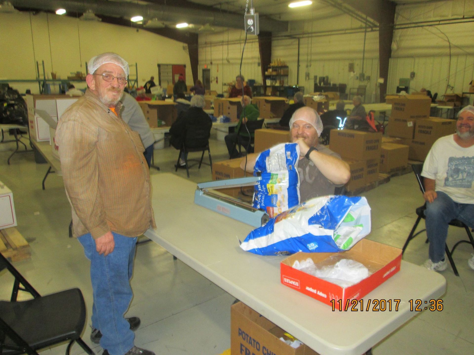 People in hairnets packing food items at tables in a warehouse. People in hairnets packing food items at tables in a warehouse.