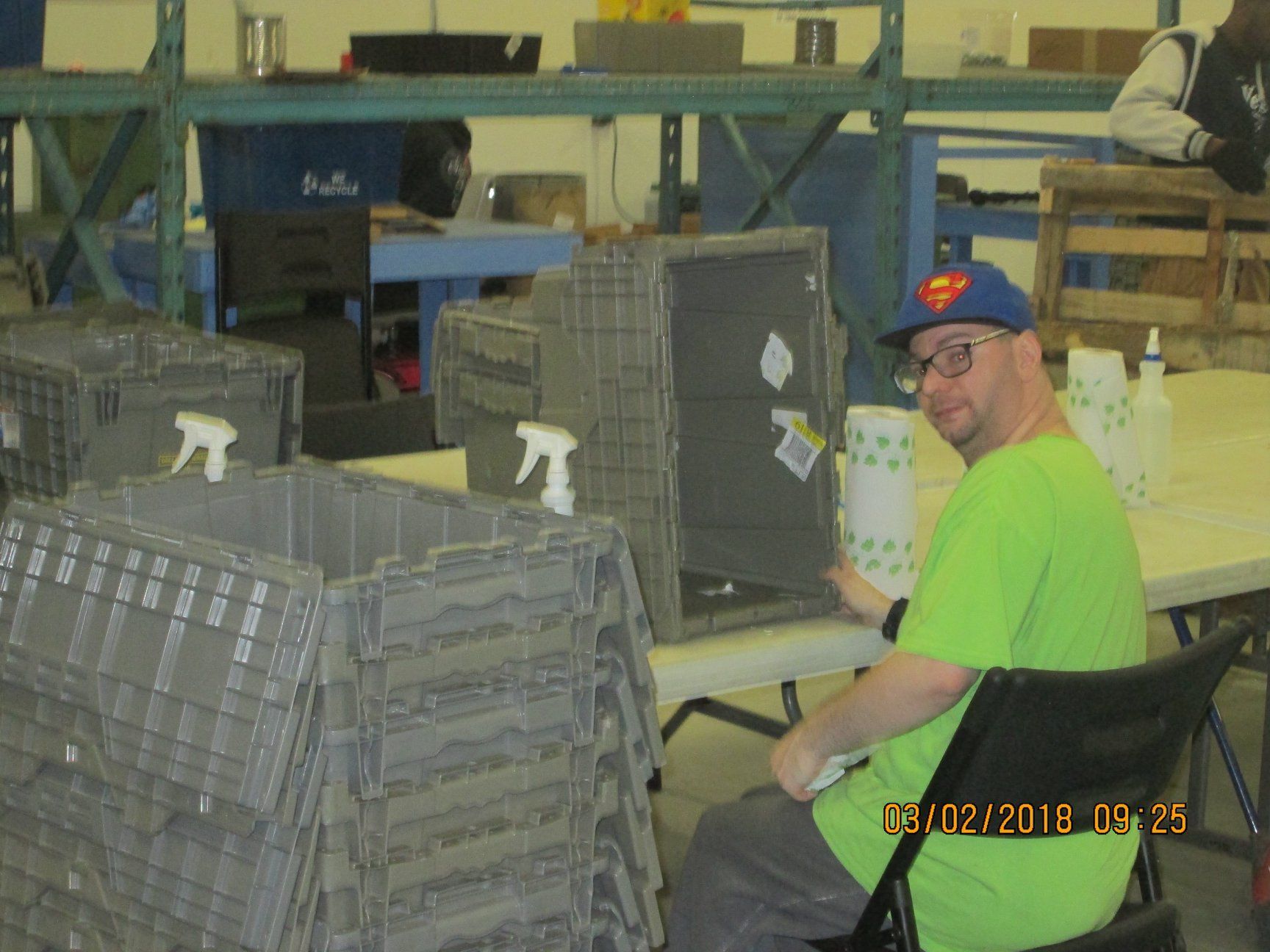 Man in green shirt, Superman hat, working at a table in a warehouse, crates stacked nearby. Man in green shirt, Superman hat, working at a table in a warehouse, crates stacked nearby.