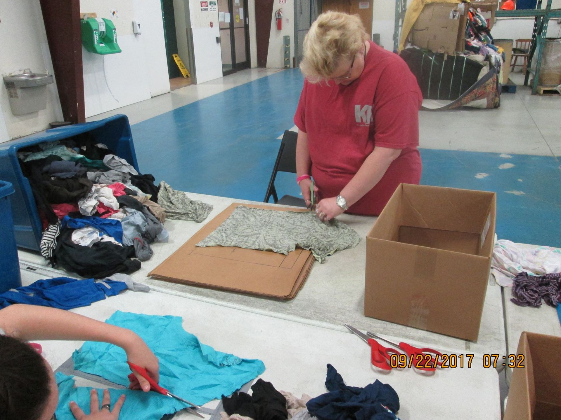 People cutting clothing at a table, likely for donation or repurposing. Blue floor, cardboard boxes, various fabrics. People cutting clothing at a table, likely for donation or repurposing. Blue floor, cardboard boxes, various fabrics.
