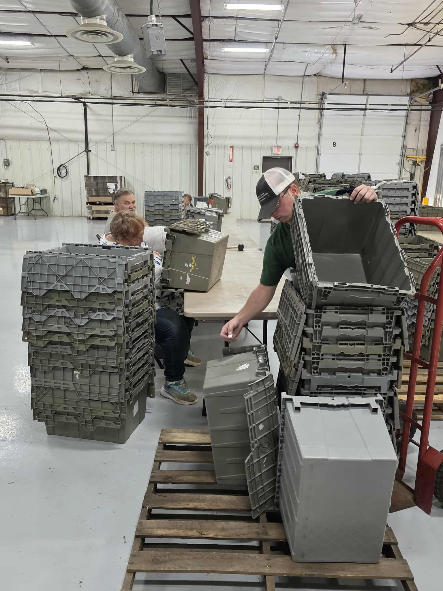Two people stack gray plastic containers in a warehouse. A man in a cap lifts one; another person sits at a table.