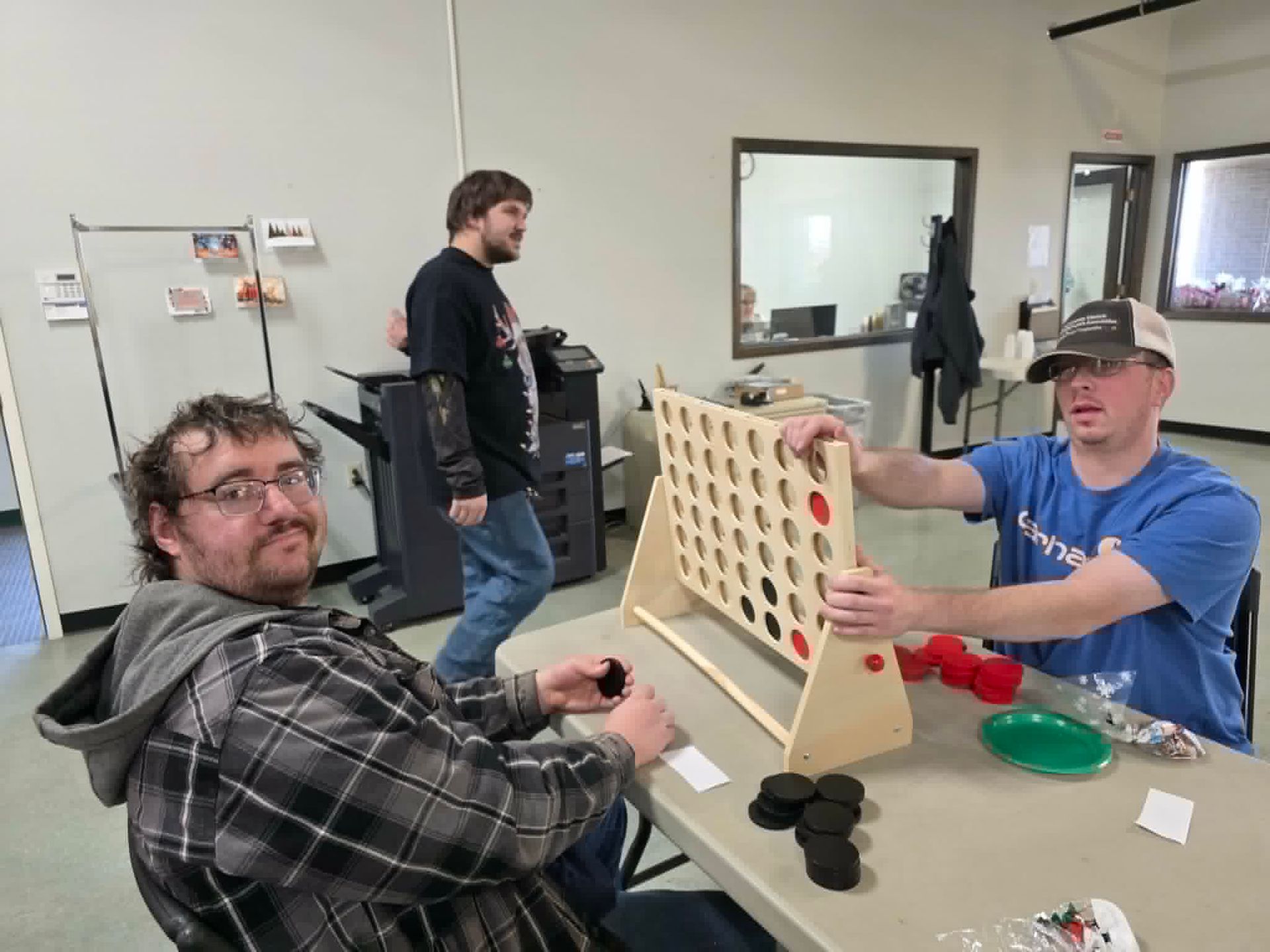 Three people playing a game of Connect Four at a table in a room. One man is looking at the camera. Three people playing a game of Connect Four at a table in a room. One man is looking at the camera.
