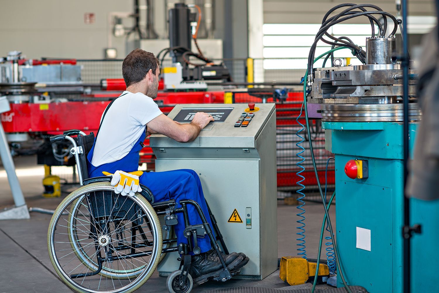Man in wheelchair operating machinery in a factory; control panel, equipment, and wires visible. Man in wheelchair operating machinery in a factory; control panel, equipment, and wires visible.