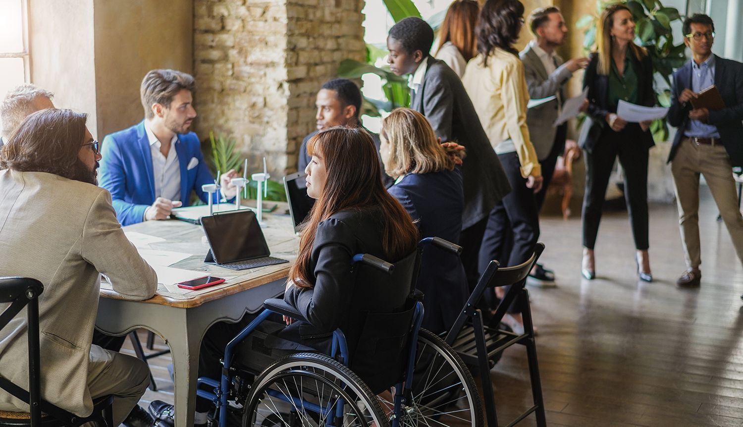 Business meeting: People at a table, one in a wheelchair, others standing and discussing in a modern office.