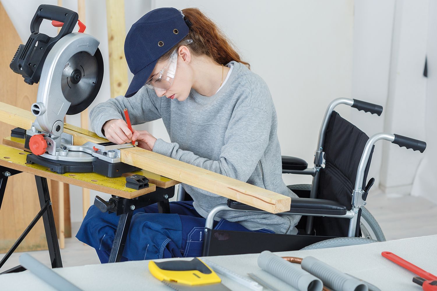 Woman in wheelchair using a miter saw to cut wood while wearing safety glasses and a cap. Woman in wheelchair using a miter saw to cut wood while wearing safety glasses and a cap.