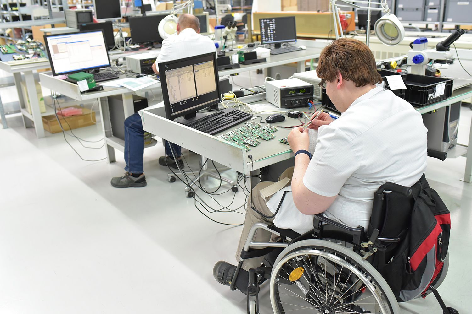 Woman in wheelchair working with electronics at a workstation. Another person works in the background. Factory setting.