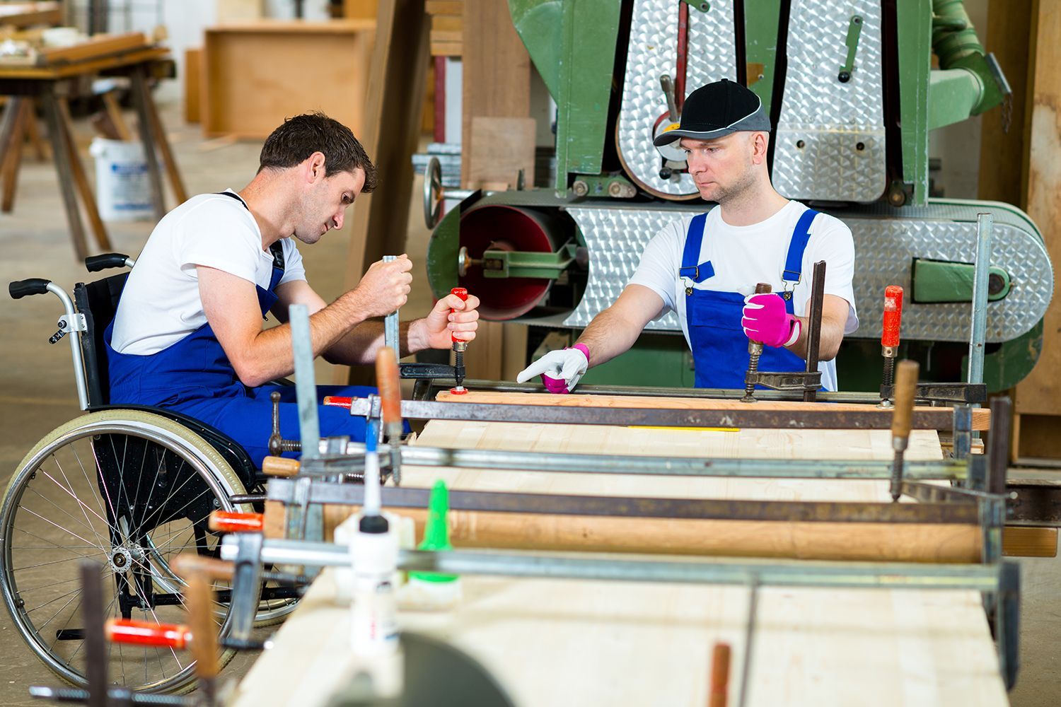 Two men working in a wood shop; one in a wheelchair, using tools at a workbench near machinery. Two men working in a wood shop; one in a wheelchair, using tools at a workbench near machinery.