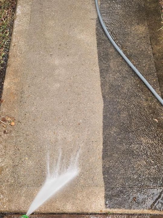 A green garden hose nozzle spraying a fan of water across a concrete driveway in front of a suburban house.