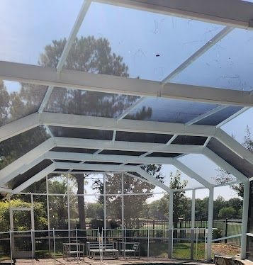 Covered outdoor patio with white beams, glass roof, and chairs beside a screened garden view