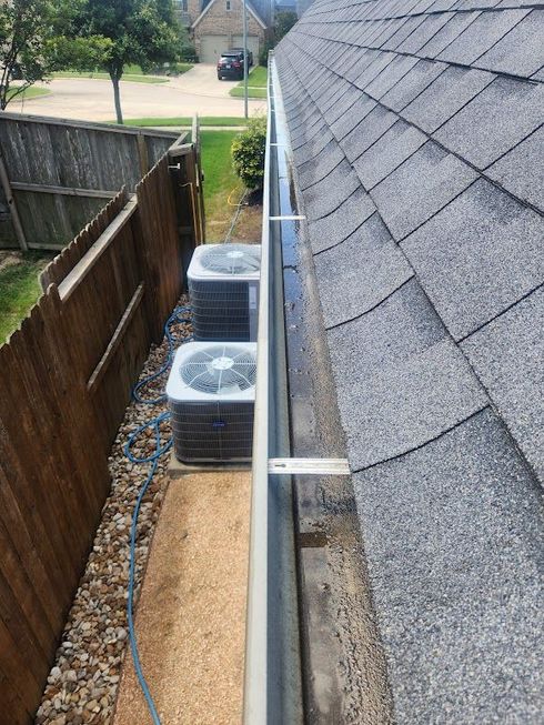 Narrow side yard with air conditioning units beside a gray shingled roof and wooden fence.