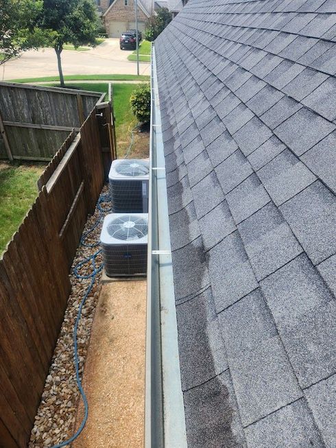Narrow side yard with two outdoor AC units beside a house, wooden fence, gravel path, and shingled roof wall.
