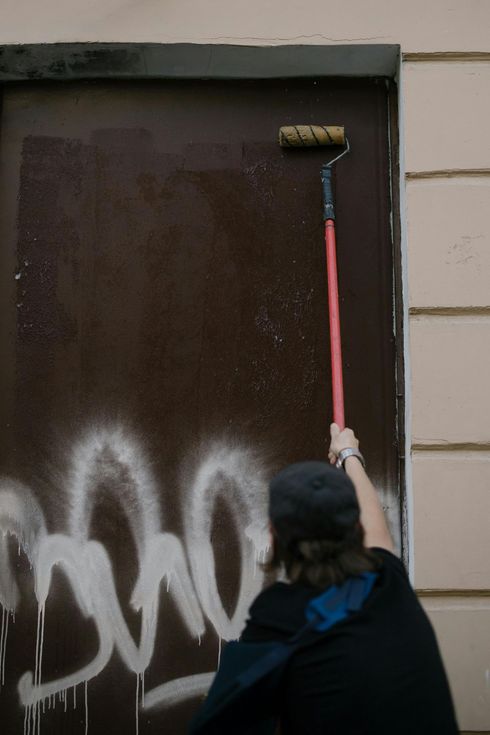 Person in dark clothing painting a brown door with a roller, with white graffiti at the bottom.