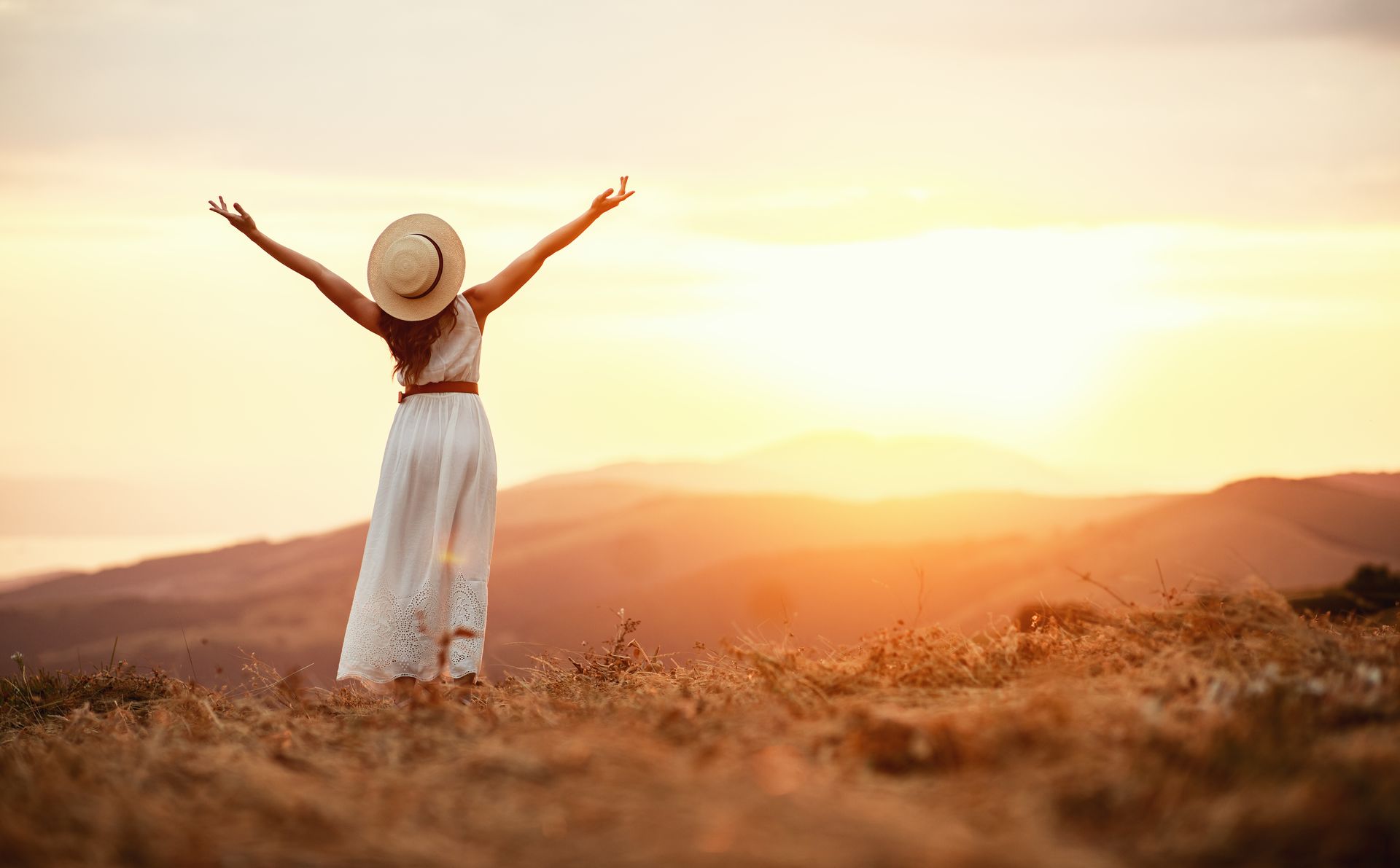 Frau mit erhobenen Armen, in Kleid und Hut, blickt auf den Sonnenuntergang über einer Berglandschaft.