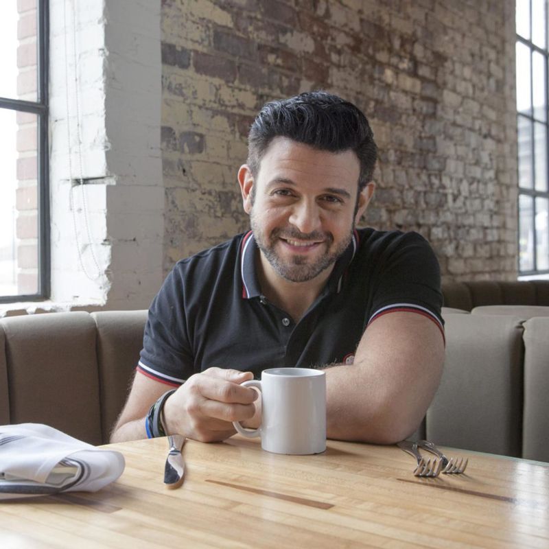 Man with dark hair smiling, holding a white mug at a restaurant table. Brick wall, natural light.