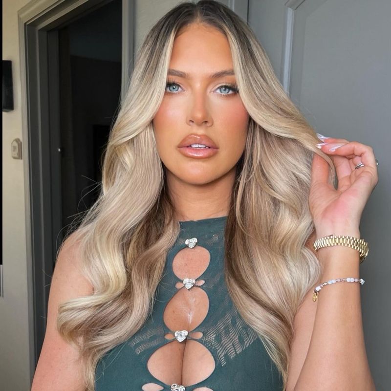 Blonde-haired woman with glam makeup and jewelry, wearing a cut-out green top, looking at the camera.