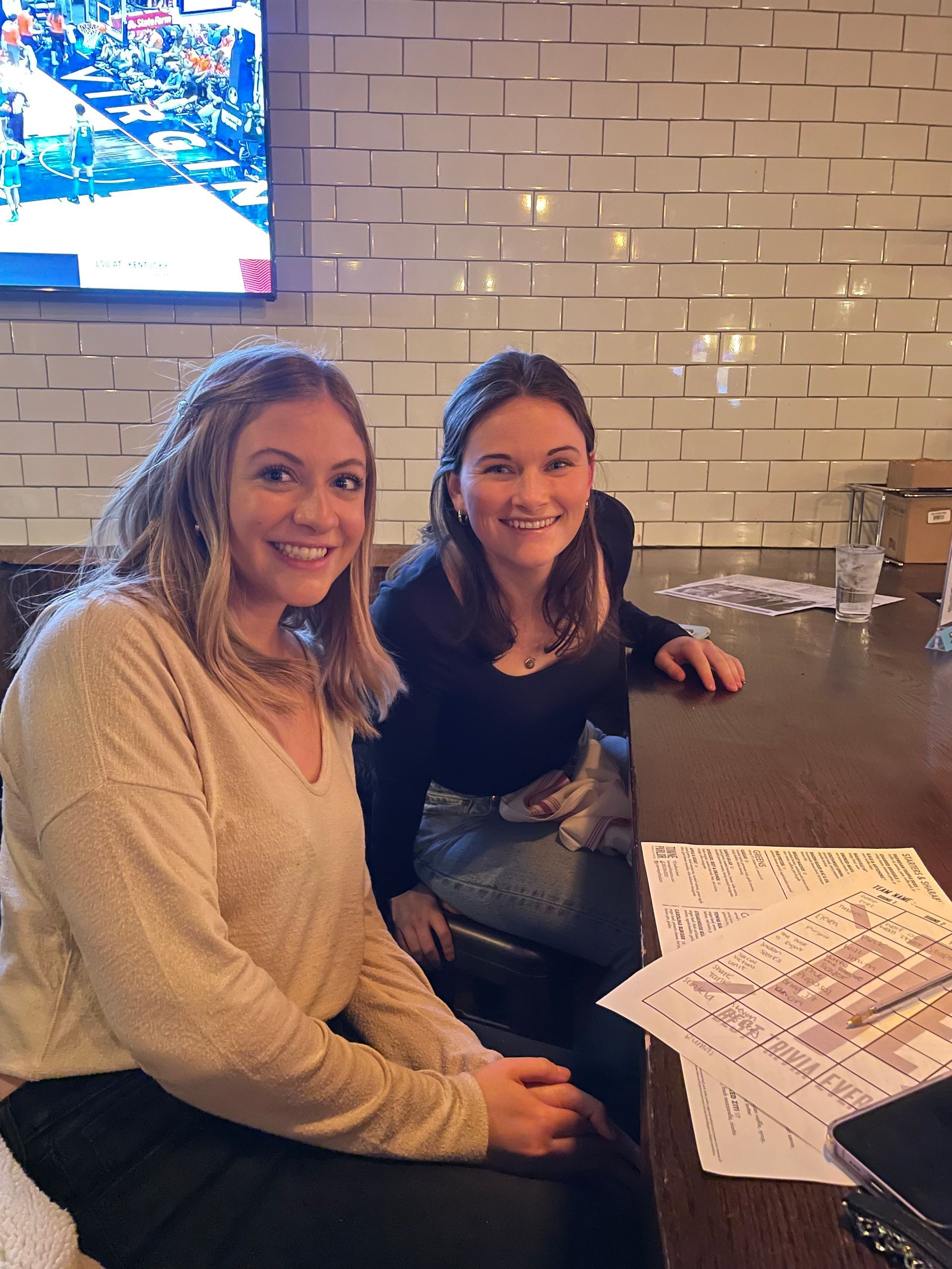 Two women smiling at a table in a bar; one in a light sweater, one in a black top.