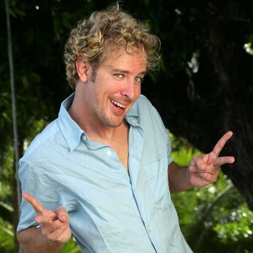 Man with curly blonde hair smiles and points fingers, wearing a blue button-down shirt outdoors.