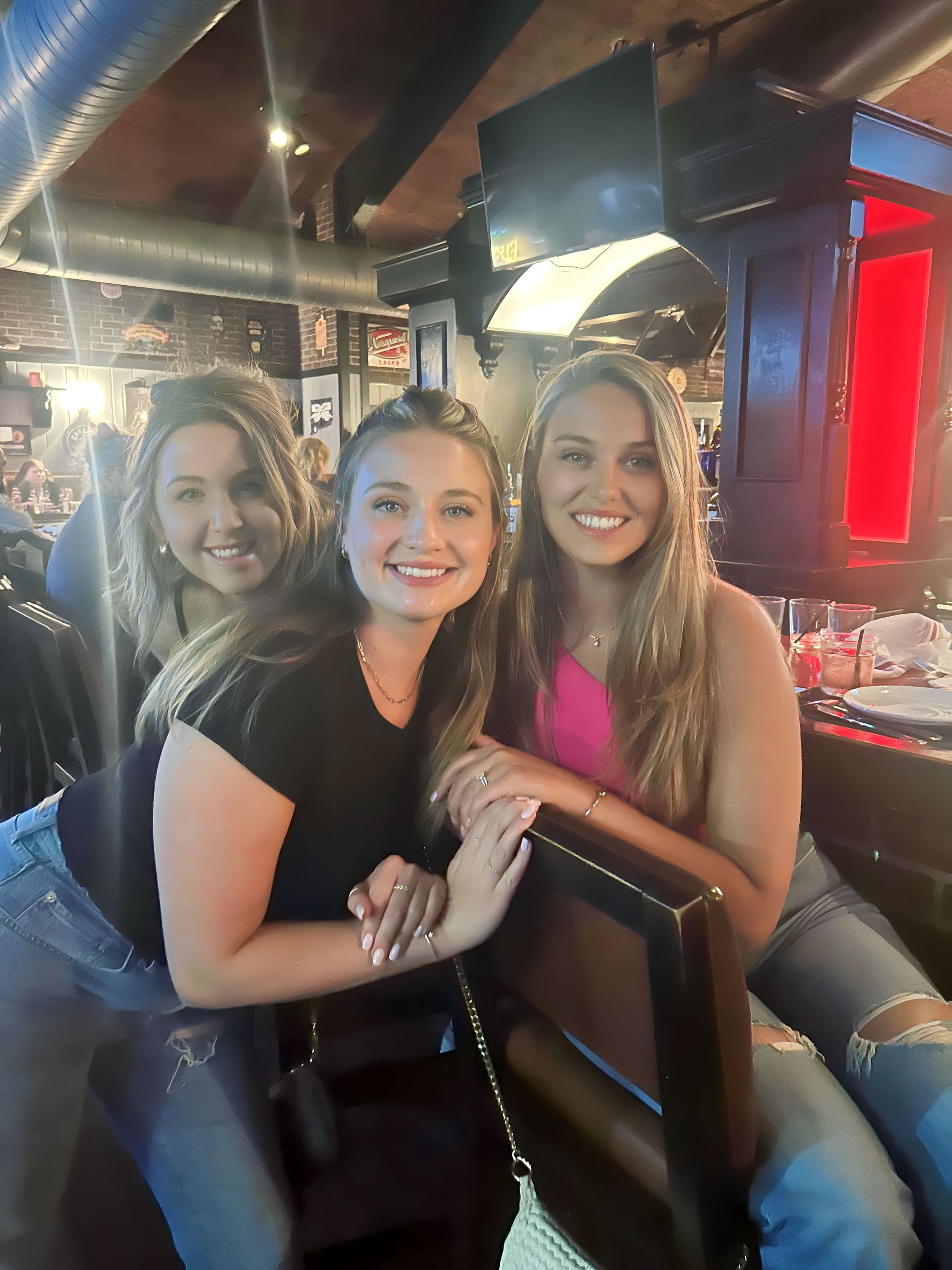 Three women smiling together in a bar. One wears pink top, another black top, all with jeans, leaning on a bar stool.