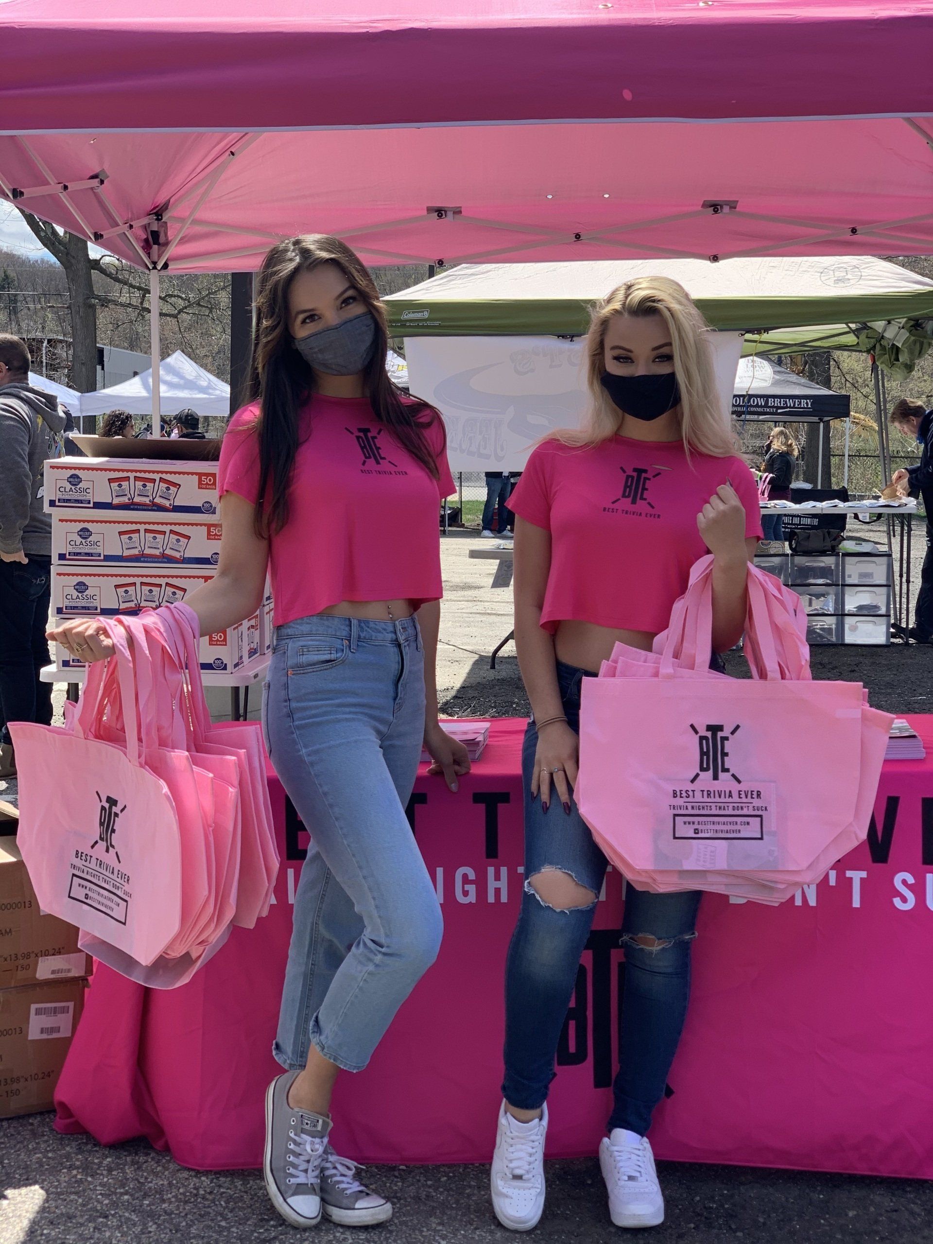 Two women in pink shirts and masks holding pink tote bags at a booth.