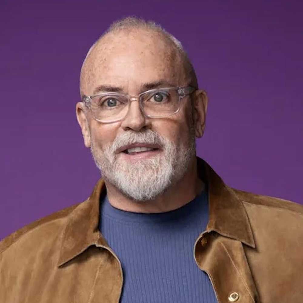 Man with gray beard and glasses, wearing a brown jacket, smiles against a purple background.